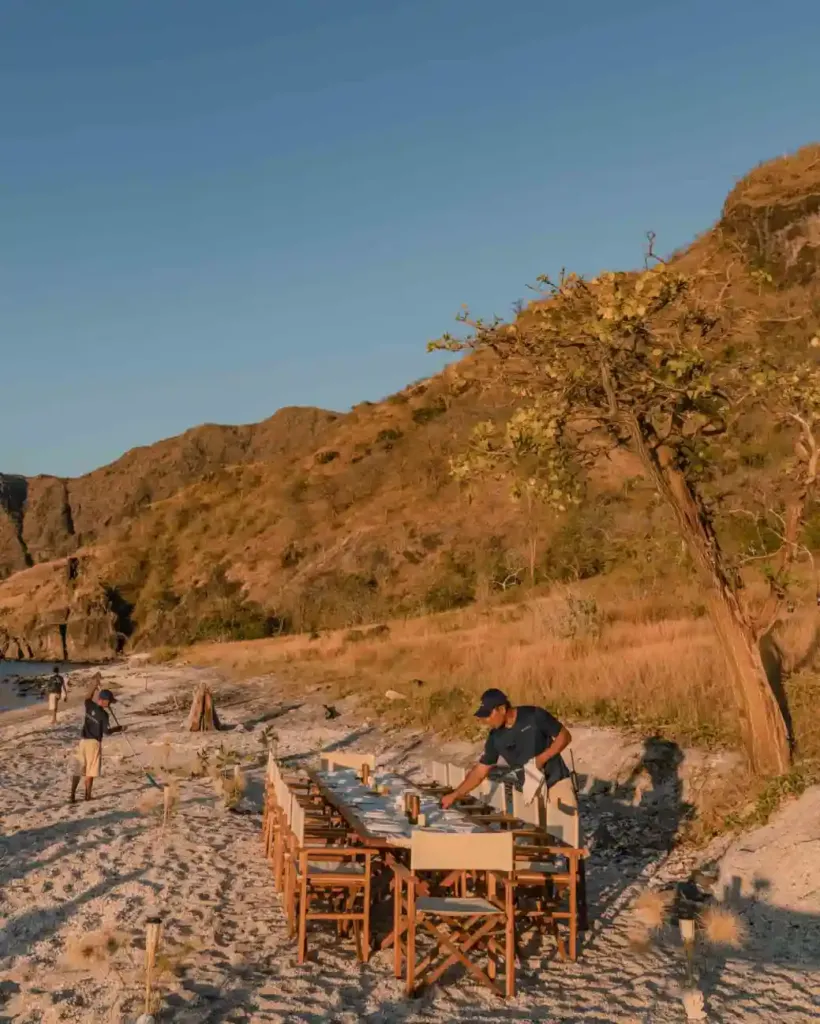 A long wooden table set on a sandy beach below dry, golden hills in warm late-afternoon light