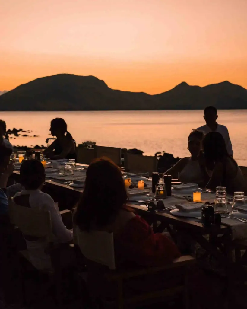 Guests at a candlelit dinner table on deck as the sky turns orange behind the islands
