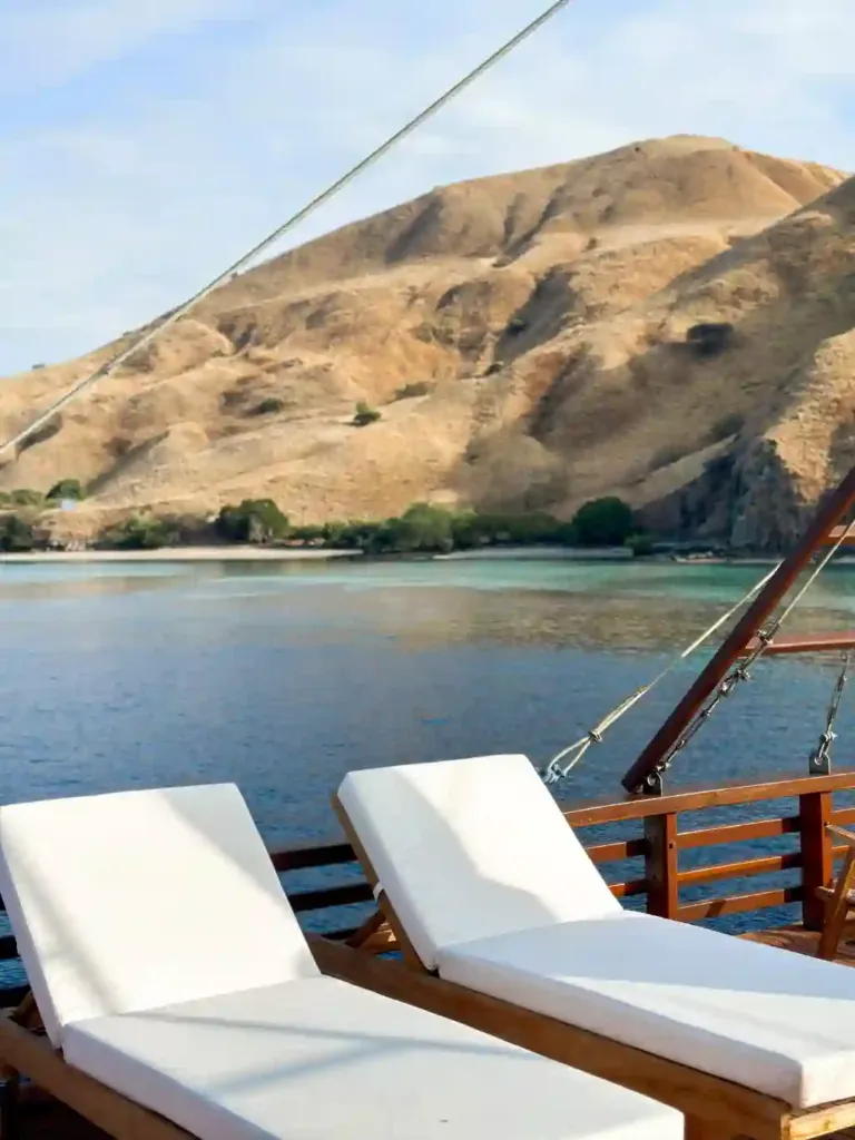 Komodo in November: white sunbeds on the deck of Neptune Cruise facing a dry, golden-brown hill above a calm bay