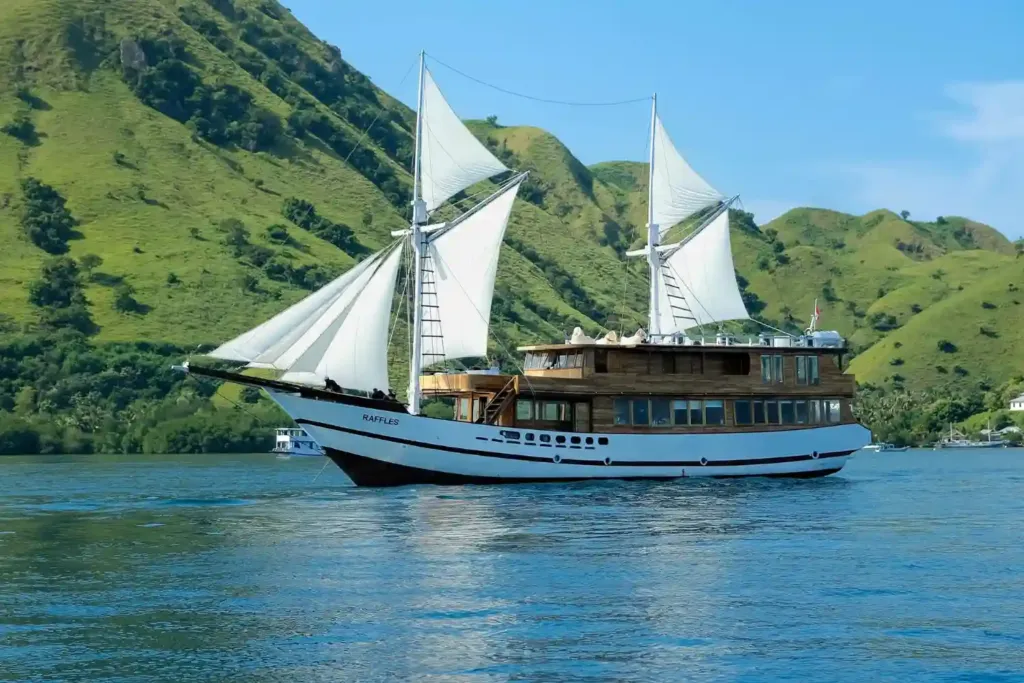 A phinisi boat under sail: Raffles Cruise in a calm bay below green hills