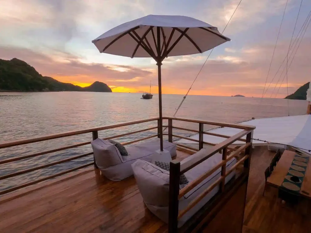 A shaded corner of a phinisi deck with umbrellas and cushions at sunset, islands on the horizon